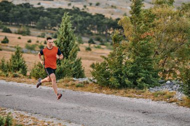 Kaslı bir erkek sporcu gün doğumunda engebeli bir dağ yolunda koşar, nefes kesen kayalık manzaralar ve doğal güzelliklerle çevrilidir.. 