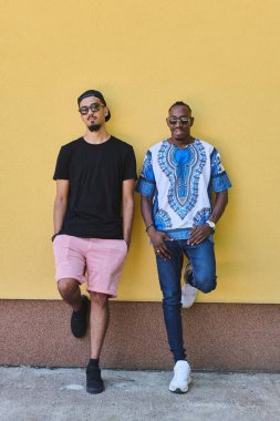 Yoldaşlık ve kültürel çeşitliliğin bir tasviri. İki erkek arkadaş, biri Sudan 'da geleneksel giyinmiş Afro-Amerikalı bir genç, canlı sarı bir duvara yaslanmış.