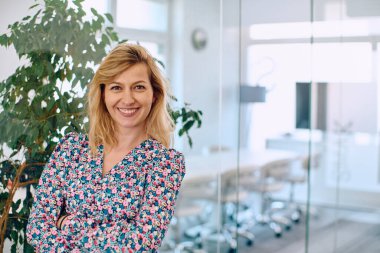 Renkli elbiseli gülümseyen bir iş kadını şirket koridorunda zarafetle duruyor. İş yerinde profesyonellik ve canlılığı somutlaştırıyor..