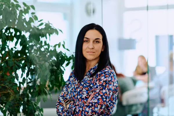 stock image A portrait captures a businesswoman with crossed arms, radiating confidence and authority amidst her bustling office environment. 