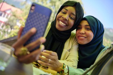 Türbanla süslenmiş iki Ortadoğulu Müslüman kadın, doğal bir ortamda otururken akıllı bir telefondan selfie çekerken bir anlık dostluk ve neşe yakalıyorlar.. 