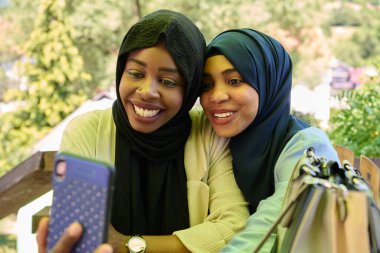 Türbanla süslenmiş iki Ortadoğulu Müslüman kadın, doğal bir ortamda otururken akıllı bir telefondan selfie çekerken bir anlık dostluk ve neşe yakalıyorlar.. 
