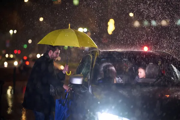 stock image A family man lovingly opens the car for his family in rainy weather, sheltering them with an umbrella.