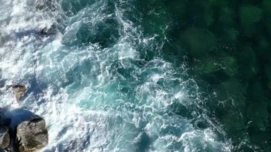 Engebeli, kayalık kıyı şeridini yıkayan turkuaz okyanus sularının hava görüntüleri. Deniz dalgaları kıyıya vuruyor. Yukarıdan görünüyor. Yüksek kalite sinematik yavaş çekim görüntüleri.