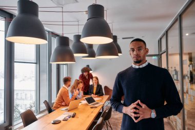 Modern bir ofis ortamında, Afrikalı bir Amerikalı işadamı, arka planda meslektaşlarıyla özgüven içinde duruyor, liderlik ve takım çalışmasını somutlaştırıyor..