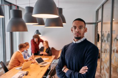 Modern bir ofis ortamında, Afrikalı bir Amerikalı işadamı, arka planda meslektaşlarıyla özgüven içinde duruyor, liderlik ve takım çalışmasını somutlaştırıyor..