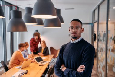 Modern bir ofis ortamında, Afrikalı bir Amerikalı işadamı, arka planda meslektaşlarıyla özgüven içinde duruyor, liderlik ve takım çalışmasını somutlaştırıyor..