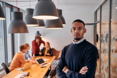 Modern bir ofis ortamında, Afrikalı bir Amerikalı işadamı, arka planda meslektaşlarıyla özgüven içinde duruyor, liderlik ve takım çalışmasını somutlaştırıyor..