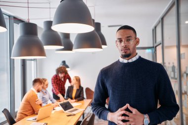 Modern bir ofis ortamında, Afrikalı bir Amerikalı işadamı, arka planda meslektaşlarıyla özgüven içinde duruyor, liderlik ve takım çalışmasını somutlaştırıyor..