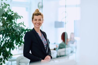 Modern bir ofiste çalışan sarışın bir iş kadınının portresi. Çalışma alanı ve arka planda meslektaşları görünüyor.. 