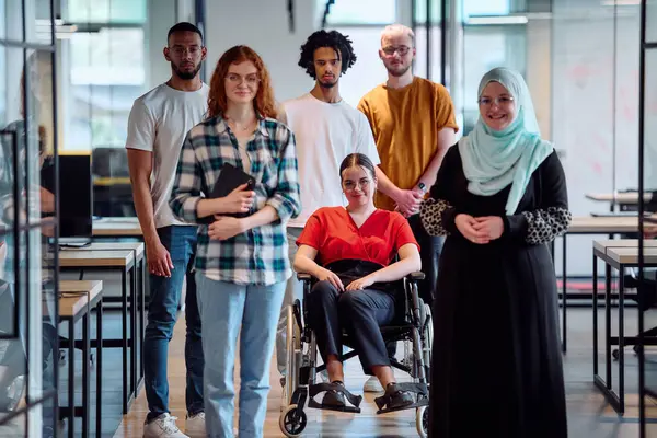 stock image A diverse group of young business people walking a corridor in the glass-enclosed office of a modern startup, including a person in a wheelchair and a woman wearing a hijab.