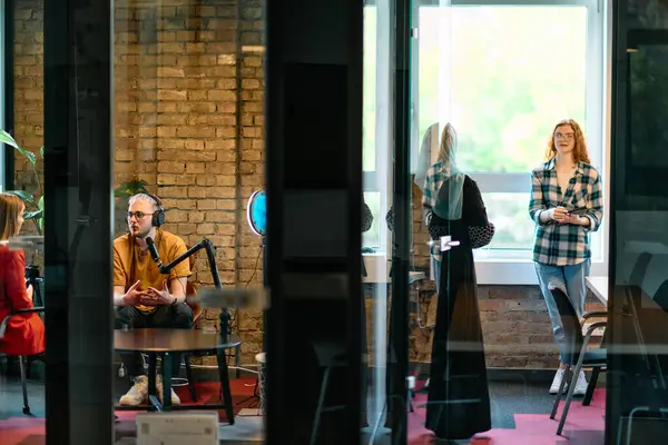 stock image A gathering of young business professionals, some seated in a glass-walled office, engage in a lively conversation and record an online podcast, embodying modern collaboration and dynamic interaction