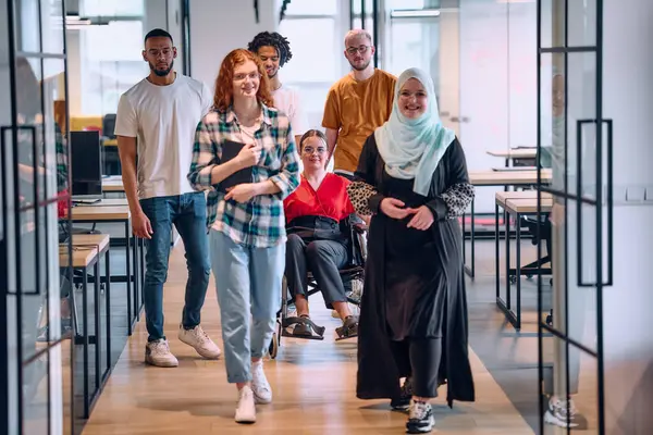  Modern bir girişimin cam kaplı ofisinde koridorda yürüyen insanlar. Tekerlekli sandalyedeki bir kişi ve tesettürlü bir kadın da dahil..