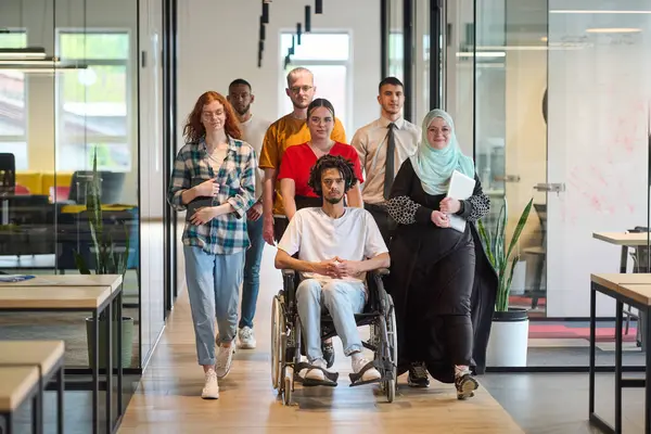 stock image A diverse group of young business people congregates within a modern startups glass-enclosed office, featuring inclusivity with a person in a wheelchair, an African American young man, and a hijab