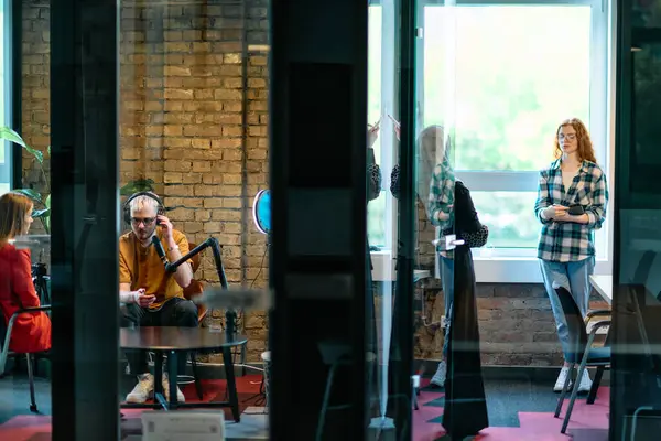 stock image A gathering of young business professionals, some seated in a glass-walled office, engage in a lively conversation and record an online podcast, embodying modern collaboration and dynamic interaction