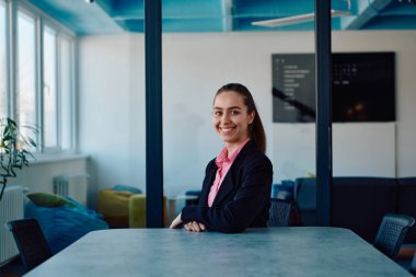 Pembe gömlekli başarılı bir bayan lider modern cam bir ofiste oturmuş kararlı bir gülümsemeyle bekliyor.