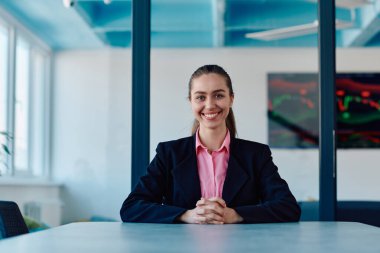 Pembe gömlekli başarılı bir bayan lider modern cam bir ofiste oturmuş kararlı bir gülümsemeyle bekliyor.