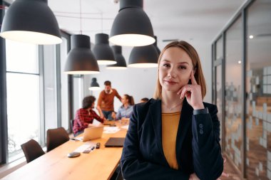 Kararlı sarışın bir iş kadını modern bir ofiste kollarını kavuşturarak duruyor. Ekibi ise özenle arka planda çalışıyor..