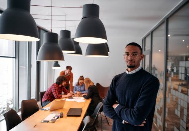 Afrika kökenli Amerikalı iş adamı modern bir ofiste çapraz silahlarla duruyor, liderlik ve güveni sembolize ederken, çeşitli meslektaşları onun arkasında duruyor, işbirliğine hazır.