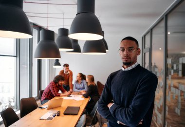 Afrika kökenli Amerikalı iş adamı modern bir ofiste çapraz silahlarla duruyor, liderlik ve güveni sembolize ederken, çeşitli meslektaşları onun arkasında duruyor, işbirliğine hazır.