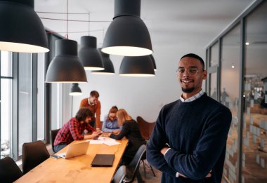 Afrika kökenli Amerikalı iş adamı modern bir ofiste çapraz silahlarla duruyor, liderlik ve güveni sembolize ederken, çeşitli meslektaşları onun arkasında duruyor, işbirliğine hazır.