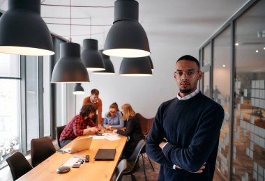 Afrika kökenli Amerikalı iş adamı modern bir ofiste çapraz silahlarla duruyor, liderlik ve güveni sembolize ederken, çeşitli meslektaşları onun arkasında duruyor, işbirliğine hazır.