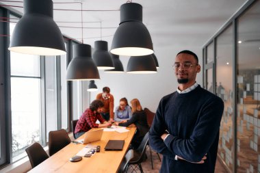 Afrika kökenli Amerikalı iş adamı modern bir ofiste çapraz silahlarla duruyor, liderlik ve güveni sembolize ederken, çeşitli meslektaşları onun arkasında duruyor, işbirliğine hazır.