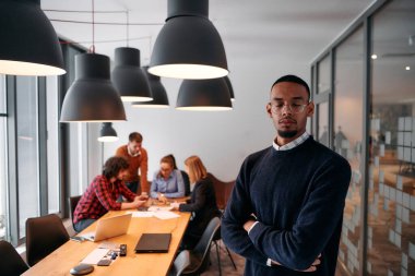 Afrika kökenli Amerikalı iş adamı modern bir ofiste çapraz silahlarla duruyor, liderlik ve güveni sembolize ederken, çeşitli meslektaşları onun arkasında duruyor, işbirliğine hazır.