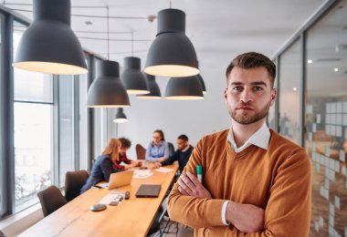Kollarını kavuşturmuş gülümseyen bir işadamı, özenli meslektaşlarıyla çevrili çağdaş bir ofiste verimli bir toplantıya liderlik ediyor.