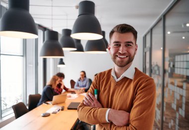 Kollarını kavuşturmuş gülümseyen bir işadamı, özenli meslektaşlarıyla çevrili çağdaş bir ofiste verimli bir toplantıya liderlik ediyor.