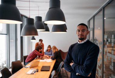 Afrika kökenli Amerikalı iş adamı modern bir ofiste çapraz silahlarla duruyor, liderlik ve güveni sembolize ederken, çeşitli meslektaşları onun arkasında duruyor, işbirliğine hazır.