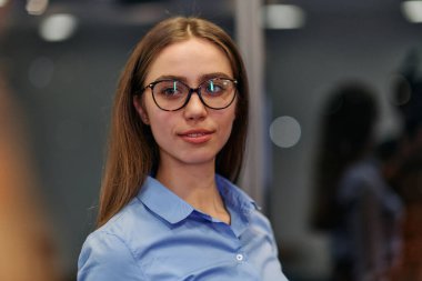 Gözlüklü ve mavi tişörtlü genç profesyonel bir iş kadını iş arkadaşlarıyla iş yerinde sohbet ediyor..