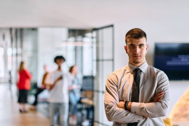 Genç bir iş adamı modern bir ofis koridorunda kollarını kavuşturarak duruyor. Kendine güven ve bir amaç duygusu yayıyor. Dinamik ve ilham verici bir varlığı somutlaştırıyor.. 