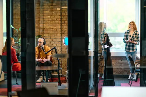 stock image A gathering of young business professionals, some seated in a glass-walled office, engage in a lively conversation and record an online podcast, embodying modern collaboration and dynamic interaction