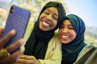 Türbanla süslenmiş iki Ortadoğulu Müslüman kadın, doğal bir ortamda otururken akıllı bir telefondan selfie çekerken bir anlık dostluk ve neşe yakalıyorlar.. 