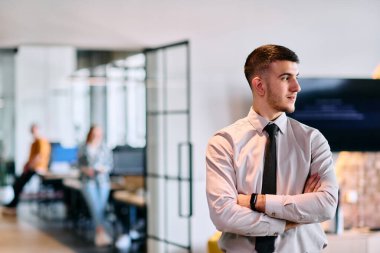 Genç bir iş adamı modern bir ofis koridorunda kollarını kavuşturarak duruyor. Kendine güven ve bir amaç duygusu yayıyor. Dinamik ve ilham verici bir varlığı somutlaştırıyor.. 