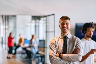 Genç bir iş adamı modern bir ofis koridorunda kollarını kavuşturarak duruyor. Kendine güven ve bir amaç duygusu yayıyor. Dinamik ve ilham verici bir varlığı somutlaştırıyor.. 