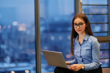 Bir iş kadını, büyük bir şirket binasının penceresinin önünde otururken dizüstü bilgisayarından faydalanıyor. Arkaplanı olarak şehrin ufuk çizgisinin manzarasını sunuyor.