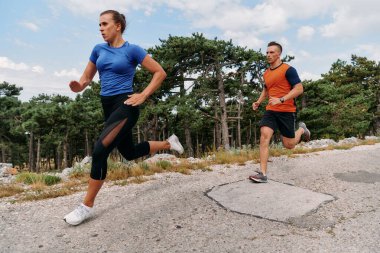 Spor kıyafetleri giymiş bir çift sabahın erken saatlerinde spor yaparken manzaralı bir yolda koşuyor, temiz havanın tadını çıkarıyor ve sağlıklı bir yaşam tarzı sürdürüyorlar..