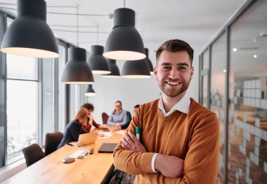 Kollarını kavuşturmuş gülümseyen bir işadamı, özenli meslektaşlarıyla çevrili çağdaş bir ofiste verimli bir toplantıya liderlik ediyor.