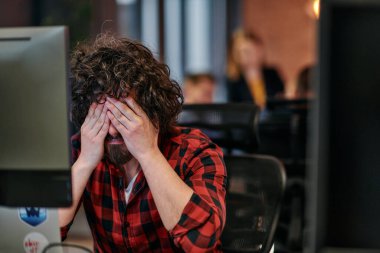 Dağınık saçlı bir programcı oturur, bilgisayar monitörüne hayal kırıklığı dolu bir bakışla bakar, teknik zorlukları yansıtır..