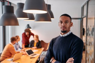 Modern bir ofis ortamında, Afrikalı bir Amerikalı işadamı, arka planda meslektaşlarıyla özgüven içinde duruyor, liderlik ve takım çalışmasını somutlaştırıyor..