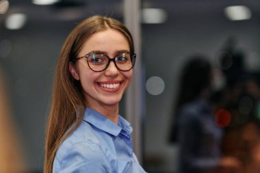 Gözlüklü ve mavi tişörtlü genç profesyonel bir iş kadını iş arkadaşlarıyla iş yerinde sohbet ediyor..
