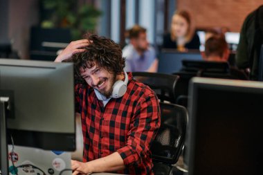 Afro saç stili ve ekose gömlekli bir programcı, modern ofis ortamındaki bir bilgisayarda özenle kodluyor.