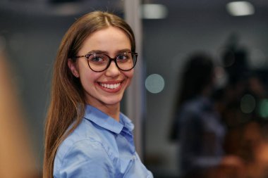 Gözlüklü ve mavi tişörtlü genç profesyonel bir iş kadını iş arkadaşlarıyla iş yerinde sohbet ediyor..