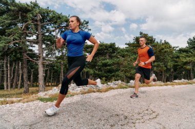 Spor kıyafetleri giymiş bir çift sabahın erken saatlerinde spor yaparken manzaralı bir yolda koşuyor, temiz havanın tadını çıkarıyor ve sağlıklı bir yaşam tarzı sürdürüyorlar..