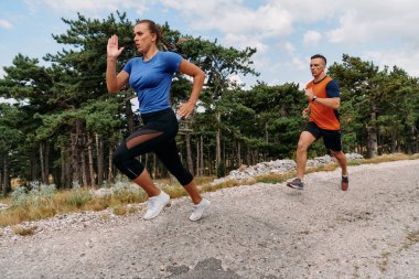 Spor kıyafetleri giymiş bir çift sabahın erken saatlerinde spor yaparken manzaralı bir yolda koşuyor, temiz havanın tadını çıkarıyor ve sağlıklı bir yaşam tarzı sürdürüyorlar..