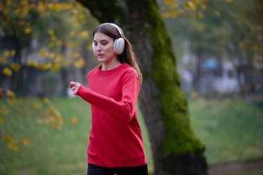 Sağlıklı yaşam tarzı, sabah koşusu. Genç ve güzel bir kadın sonbahar parkında koşuyor ve akıllı telefonuyla kulaklıkla müzik dinliyor. Kırmızı bir spor gömlek giyiyor.. 