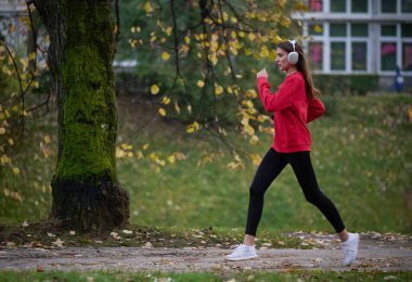 Sağlıklı yaşam tarzı, sabah koşusu. Genç ve güzel bir kadın sonbahar parkında koşuyor ve akıllı telefonuyla kulaklıkla müzik dinliyor. Kırmızı bir spor gömlek giyiyor.. 