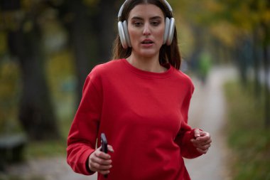 Sağlıklı yaşam tarzı, sabah koşusu. Genç ve güzel bir kadın sonbahar parkında koşuyor ve akıllı telefonuyla kulaklıkla müzik dinliyor. Kırmızı bir spor gömlek giyiyor.. 